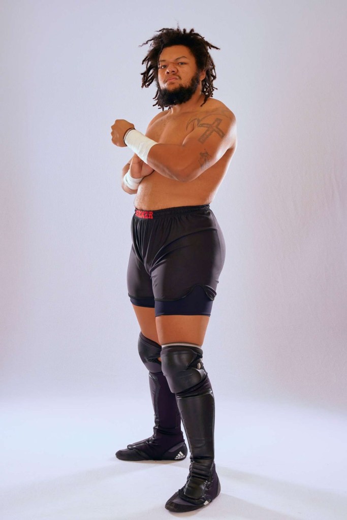 Troy Parker stands ready to battle, wearing black shorts, black shoes/shin guards, black knee pads, wrists are taped. This is a standalone portrait. 