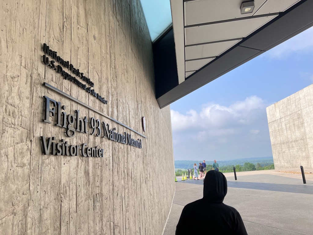 A concrete wall states Flight 93 National Memorial Visitor Center. Beyond the textured wall is a large open field. 