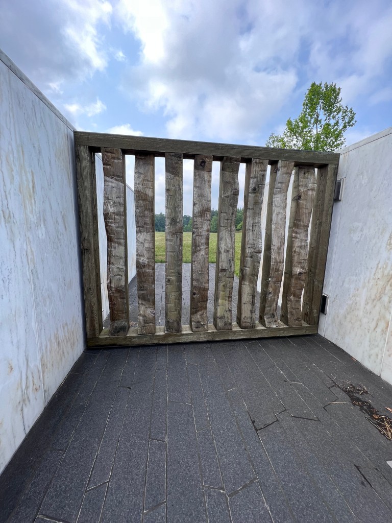 A large wooden gate made of distinct hemlock beams is closed but leads to the crash site beyond the "Wall of Names" and black granite path.