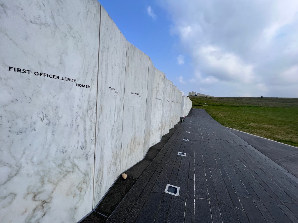Polished white marble panels have the names of crew and passengers etched into each panel. The wall extends along the black granite walkway. 