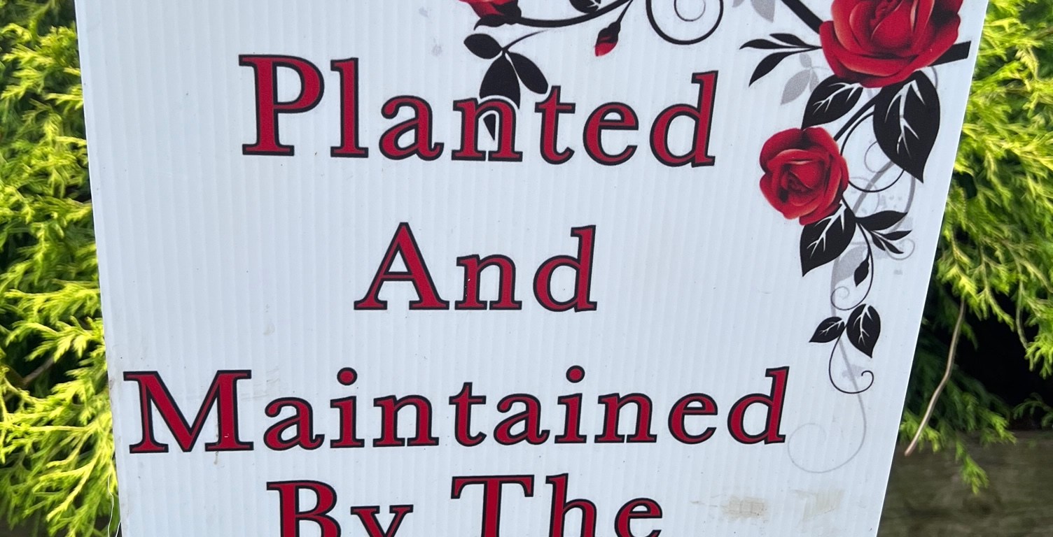 A white sign in a flower bed states "Planted And Maintained By The Tu-Endie-Wei Garden Club.