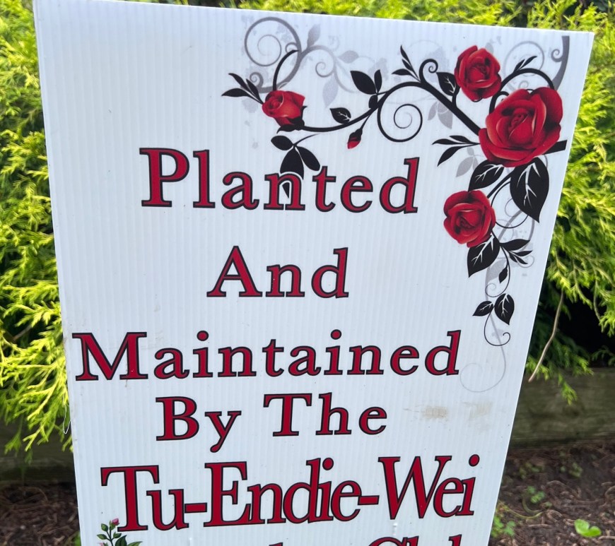 A white sign in a flower bed states "Planted And Maintained By The Tu-Endie-Wei Garden Club.