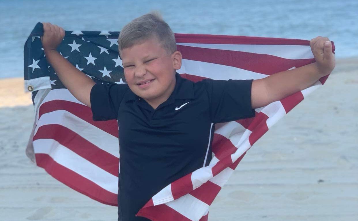 A young boy stands on the beach, holding an American Flag. The boy is Bentely Neal, who will be the grand parade marshal.
