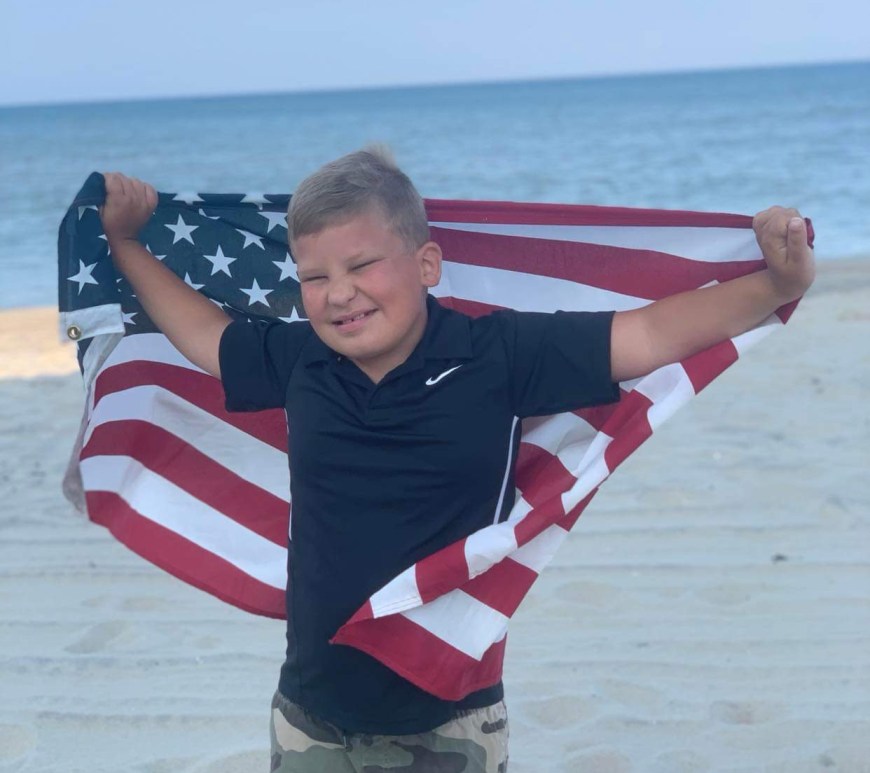 A young boy stands on the beach, holding an American Flag. The boy is Bentely Neal, who will be the grand parade marshal.