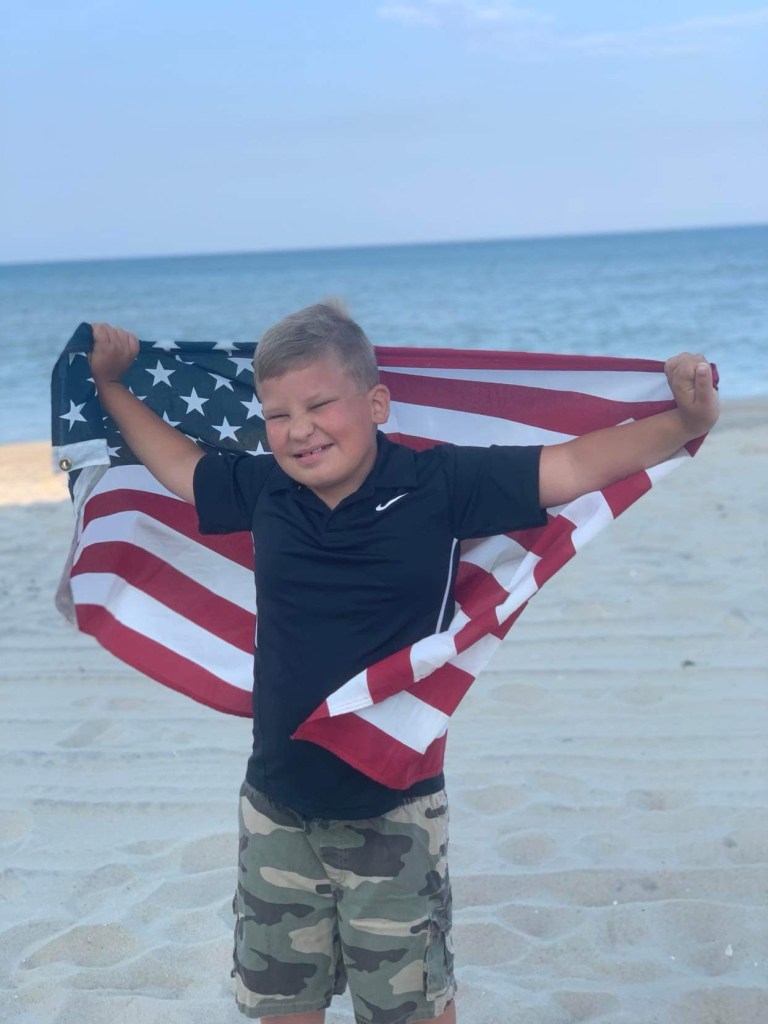 A young boy stands on the beach, holding an American Flag. The boy is Bentely Neal, the grand parade marshal for the Town of Mason’s Independence Day Parade. 