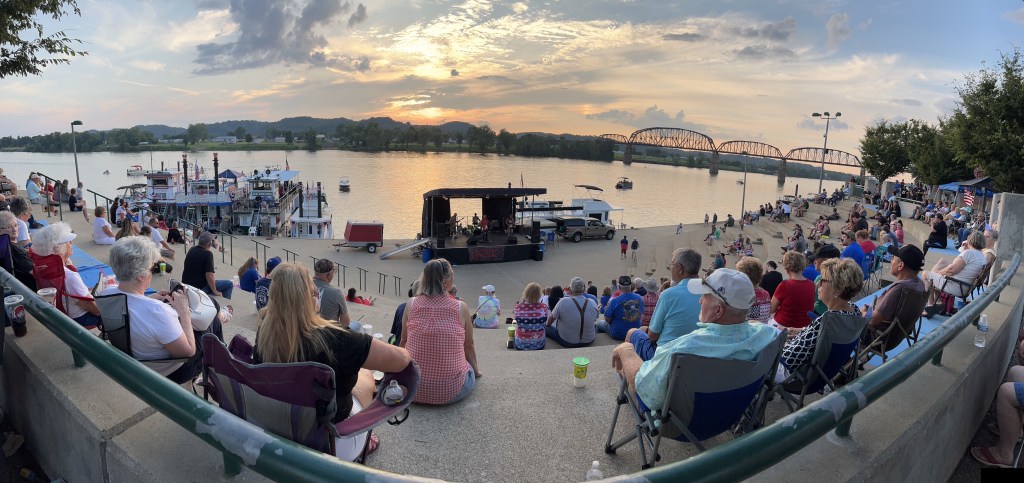 A wide shot of people filling the concrete amphitheater, listening to a band, with some sternwheelers parked along the Ohio River at sunset.