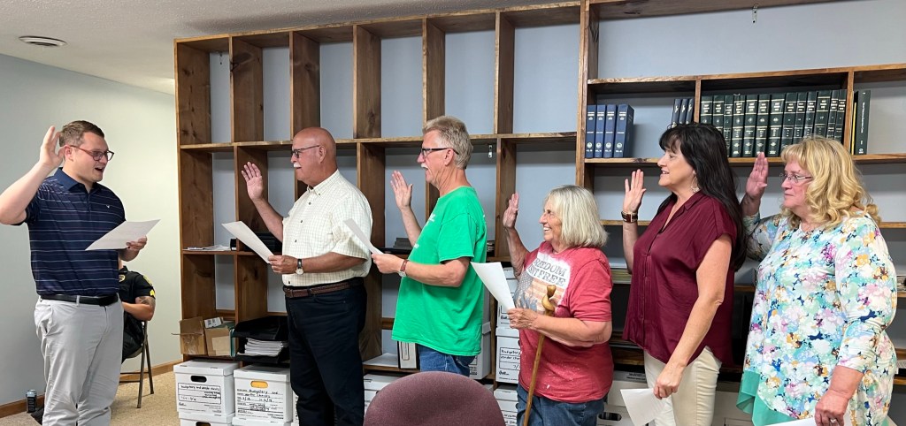 The mayor stands with his hand raised, delivering the oath of office to five council members, also with raised hands.