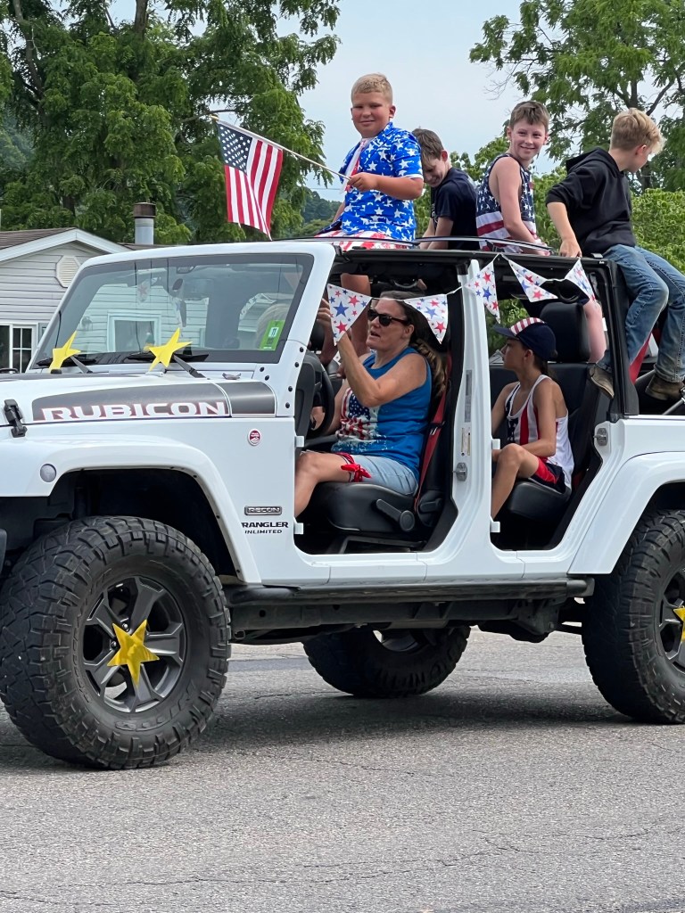 A jeep full of children smile in the parade, with the grand marshal dressed in patriotic gear. 