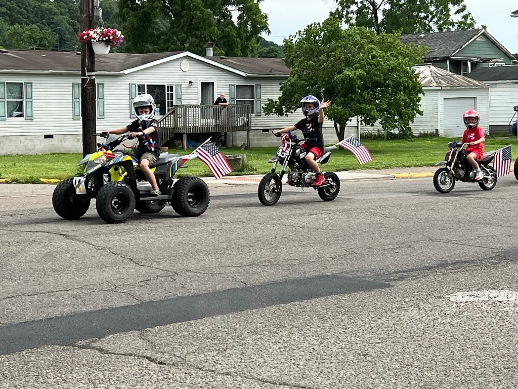 Children on dirt bikes and one on an ATV wave from the parade route.