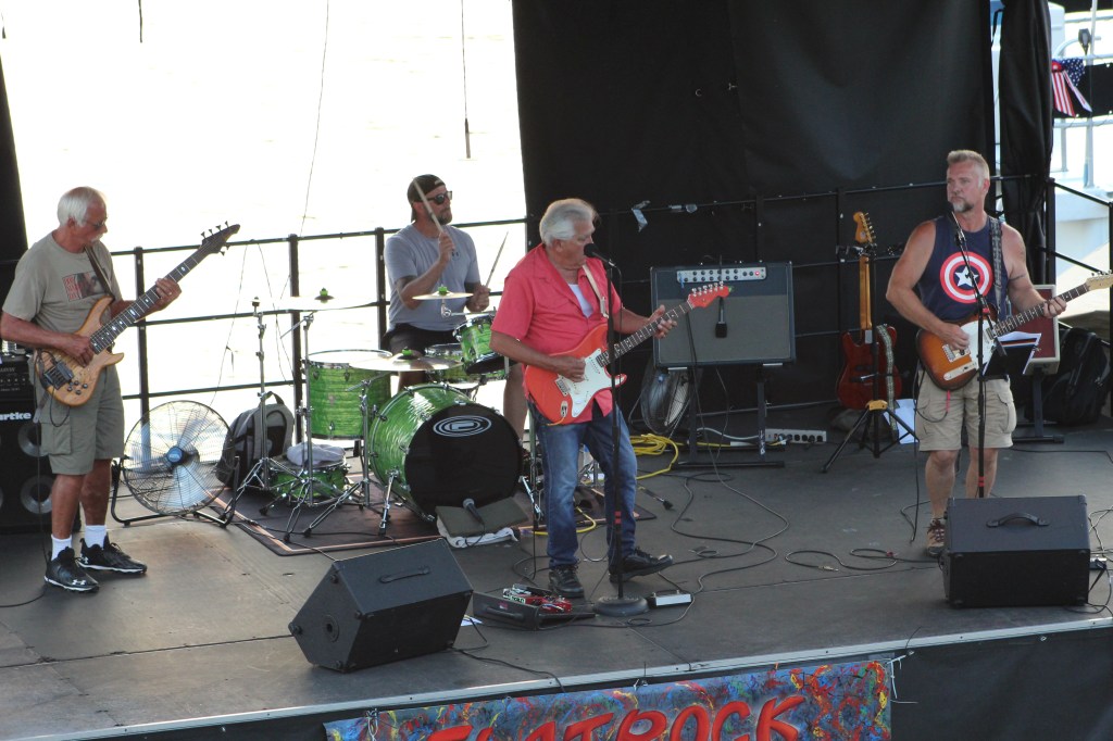 A four-piece band performs on a black  stage along the Ohio River. 