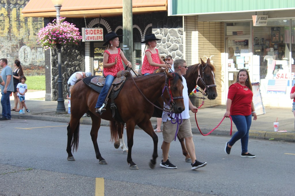 Twin girls, dressed alike, ride two horses along the parade route. 