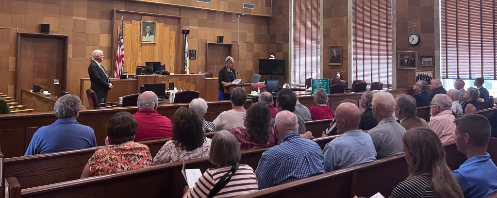 Rows of people are seated in a courtroom. One woman is speaking at a podium. A man is standing to her side.