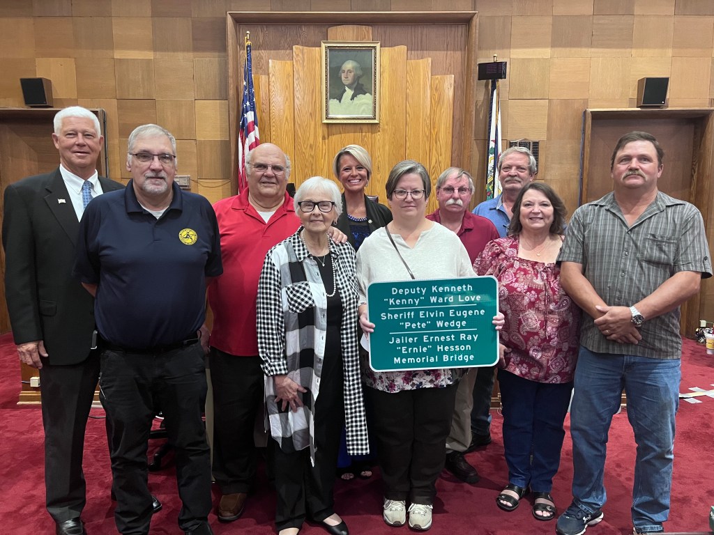 Several people are standing in a courtroom, with family members holding a sign in honor of their loved one.