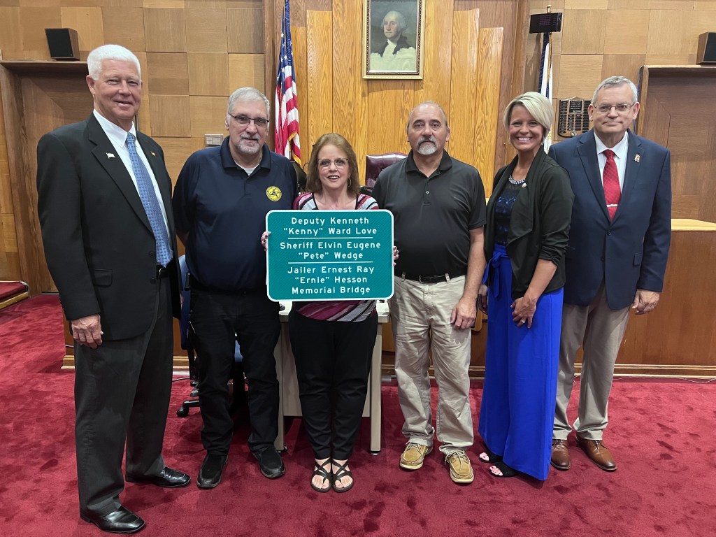 Several people are standing in a courtroom, with family members holding a sign in honor of their loved one.