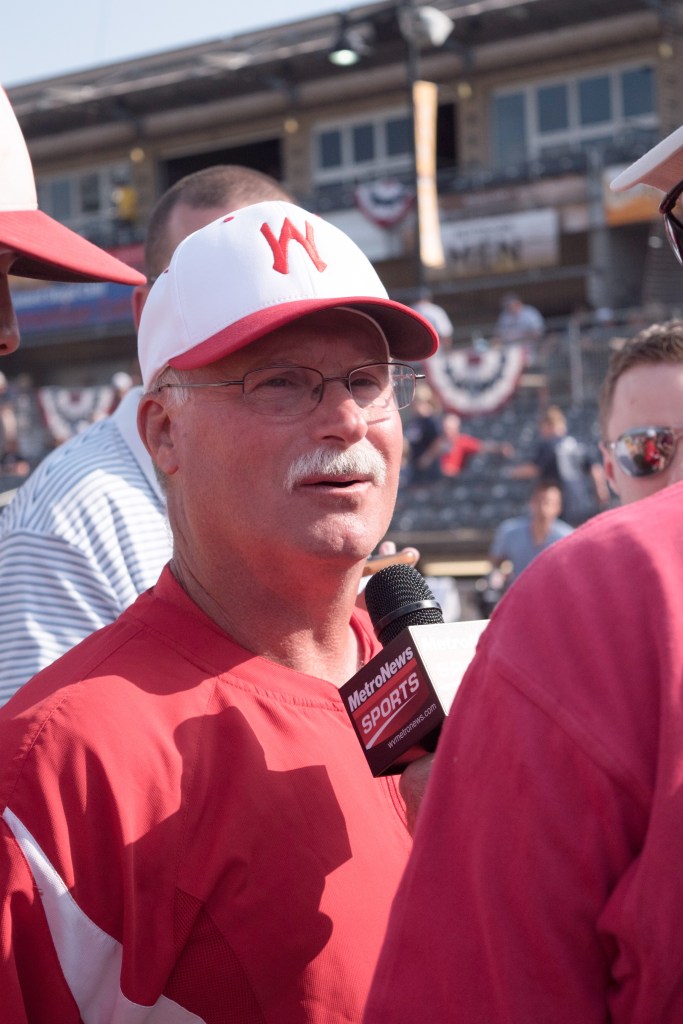 A baseball coach stands in uniform being interviewed. 