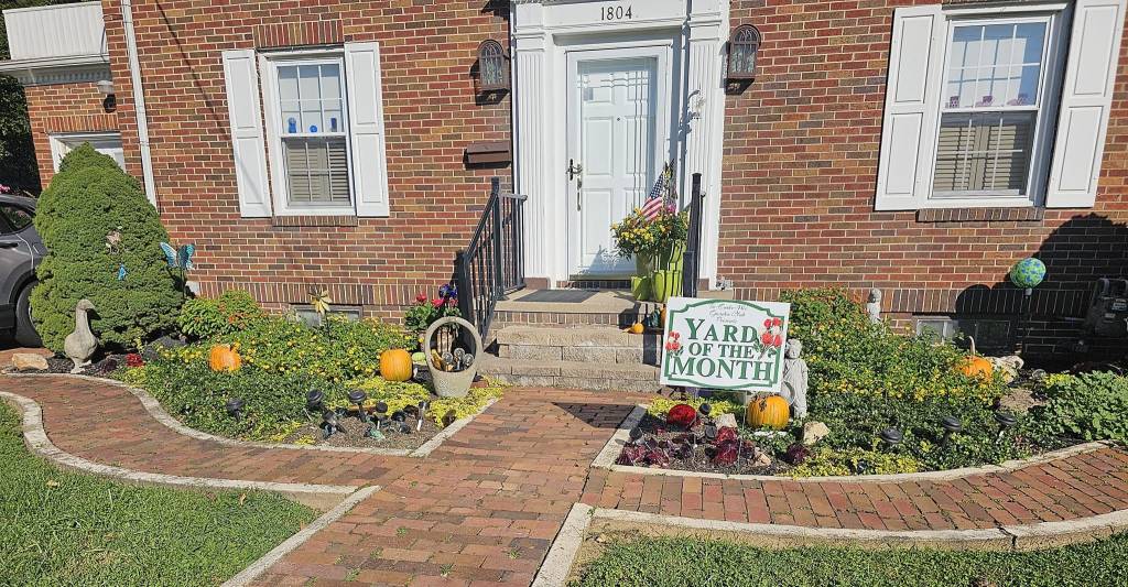 Pictured with a "Yard of the Month" sign, is a decorated yard, with flowers and landscaping surrounding a home. 