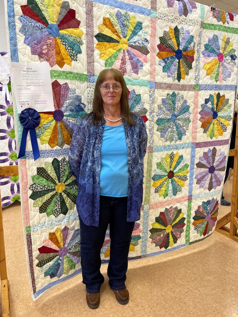 A woman stands with a large Dresden Plate quilt, with a blue riboon on it. The quilt squares all have colorful color wheels in the center of squares. 