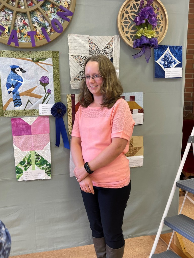 A young woman stands next to a quilt square with a blue jay stitched on it. A blue ribbon is attached. 