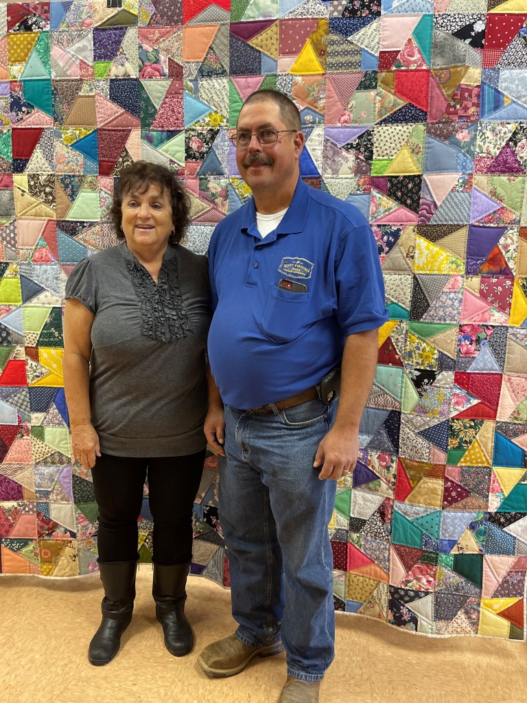 A man and woman stand in front of a large quilt that takes up an entire wall, made of several colorful squares and patterns. 