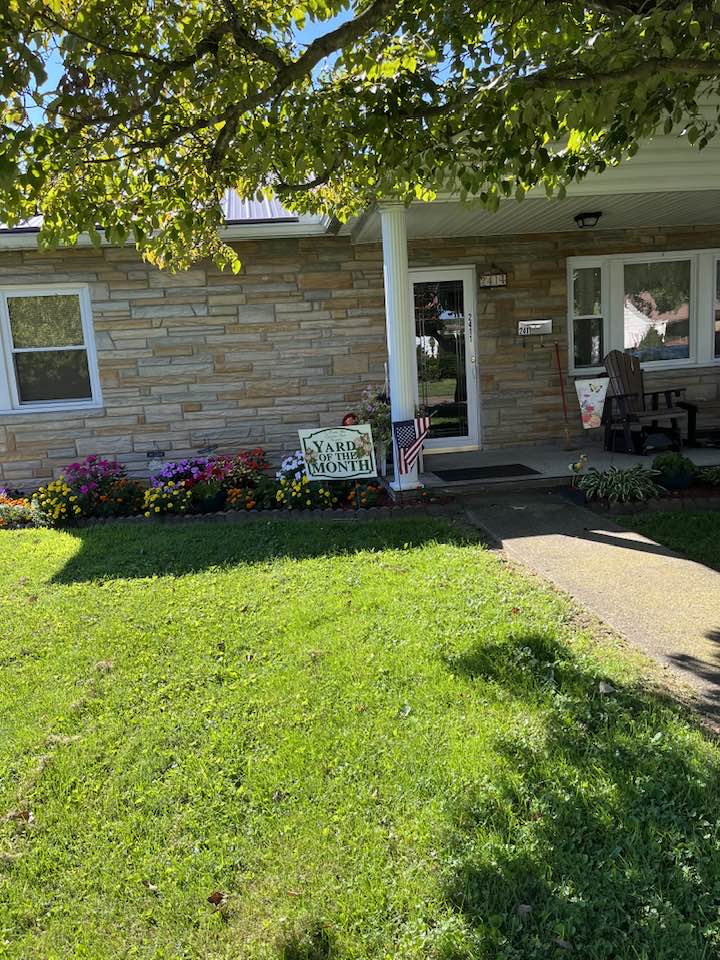 Pictured with a "Yard of the Month" sign, is a decorated yard, with flowers and landscaping surrounding a home. 