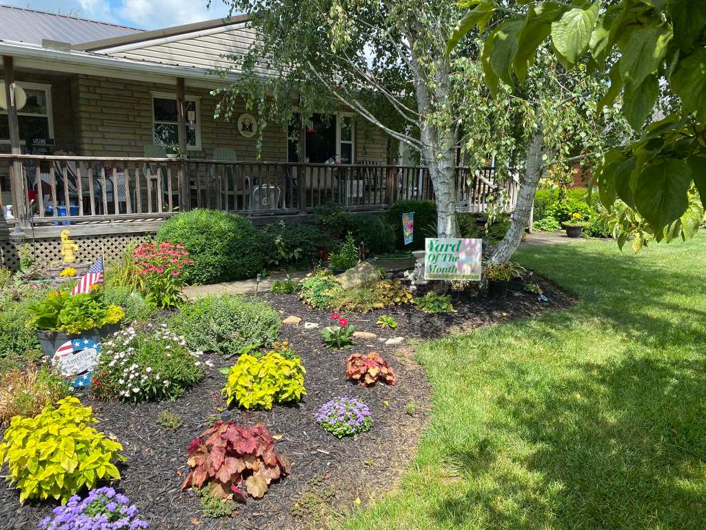 Pictured with a "Yard of the Month" sign, is a decorated yard, with flowers and landscaping surrounding a home. 
