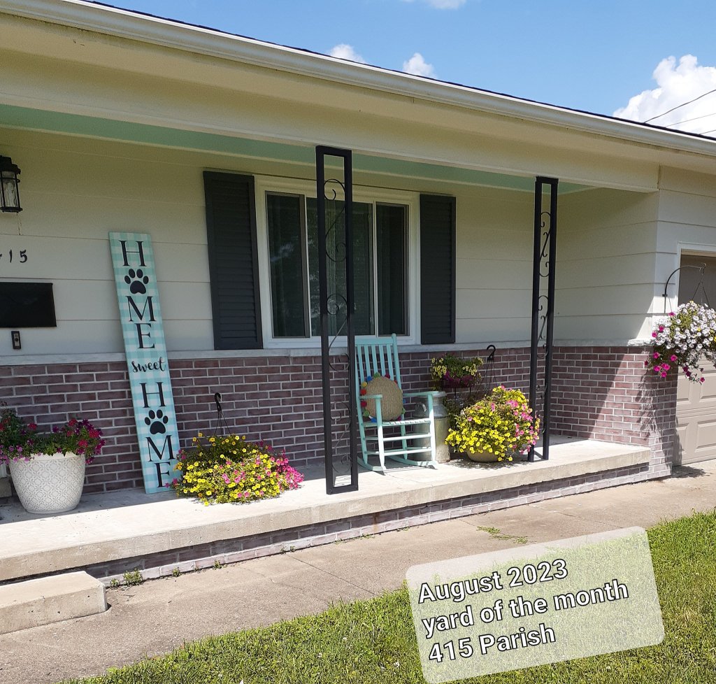 Pictured with a "Yard of the Month" sign, is a decorated yard, with flowers and landscaping surrounding a home. 