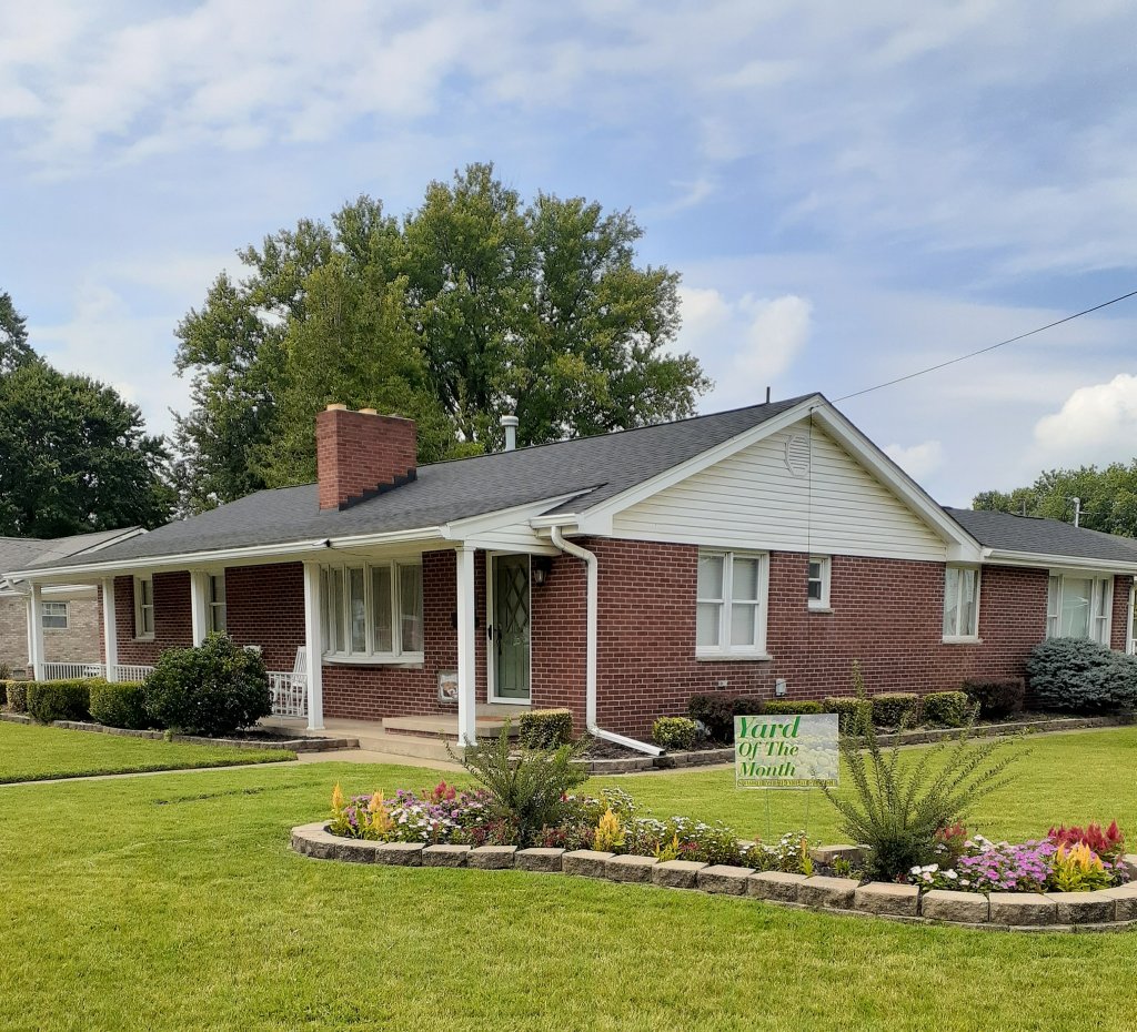 Pictured with a "Yard of the Month" sign, is a decorated yard, with flowers and landscaping surrounding a home. 
