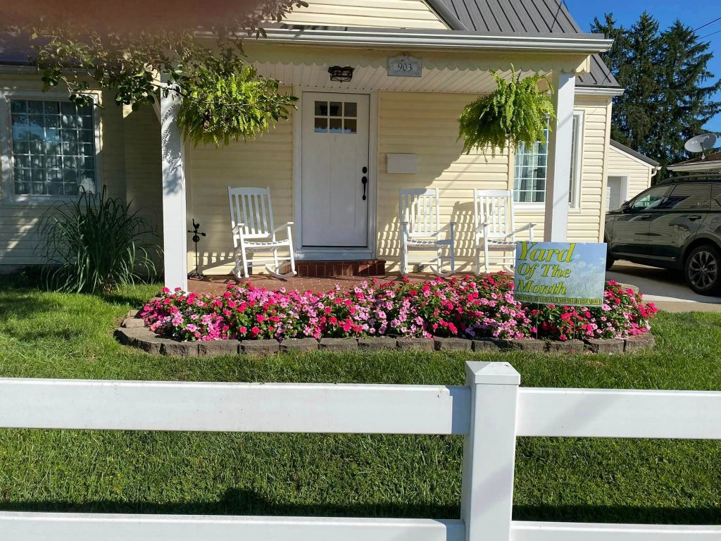 Pictured with a "Yard of the Month" sign, is a decorated yard, with flowers and landscaping surrounding a home. 