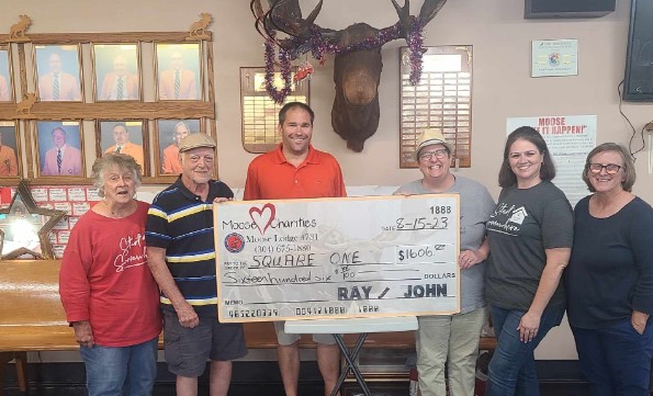 A group of six people stand behind a large check, made out to a local charity. 