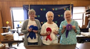 Three ladies stand holding rosettes in made of blue and red material. 