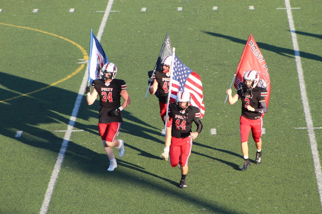Four football players enter the field, each carrying a flag representing their state, school, program and U.S. Flag. 