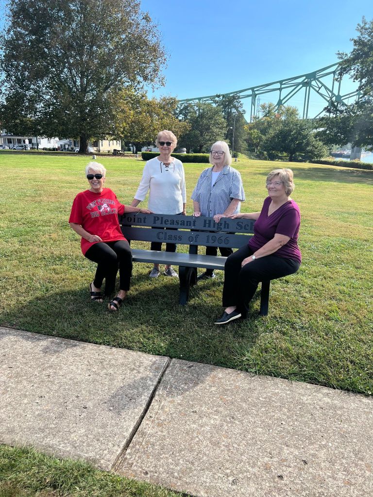 Four classmates sit on a gray bench with the words "Point Pleasant High School Class of 1966" at the park where the bench was donated. 