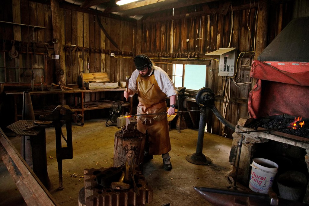 A blacksmith stands with an instrument to mold hot metal in a blacksmith shop. Hot coals are in the background.