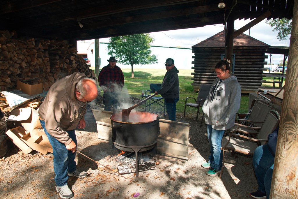 A group of four people stand around a large kettle over an open fire, stirring apple butter.