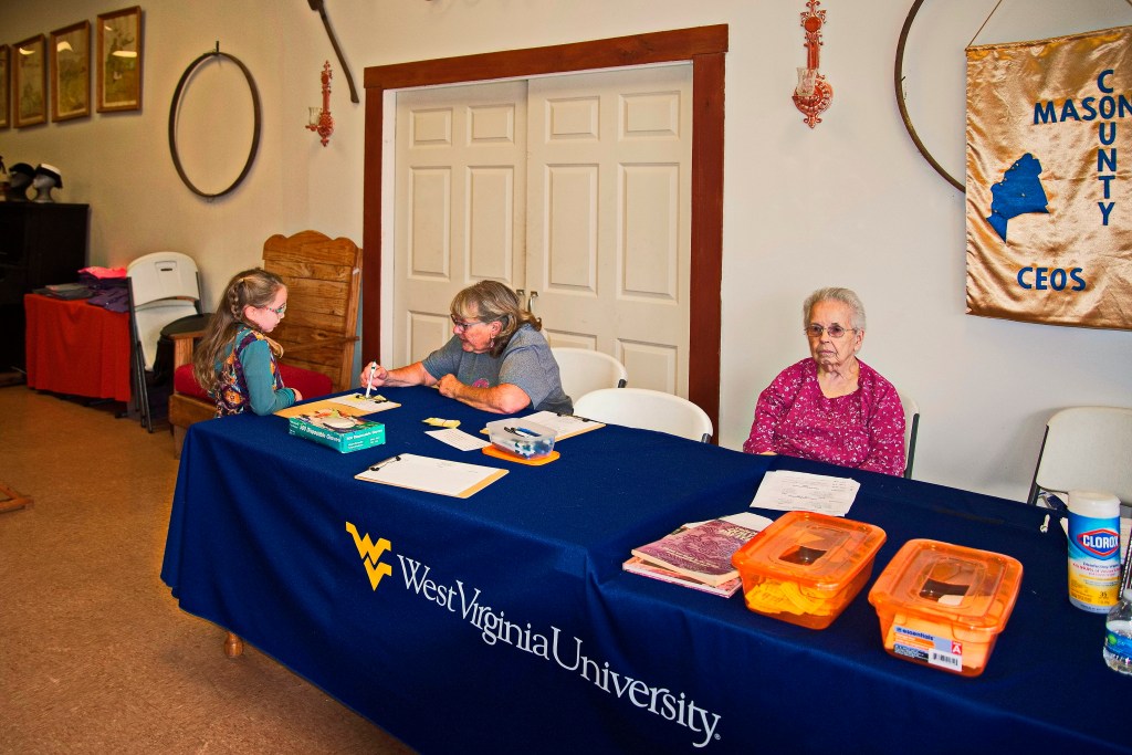 Two women sit at a registration table for a quilt show. A child asks one of the women a question. 