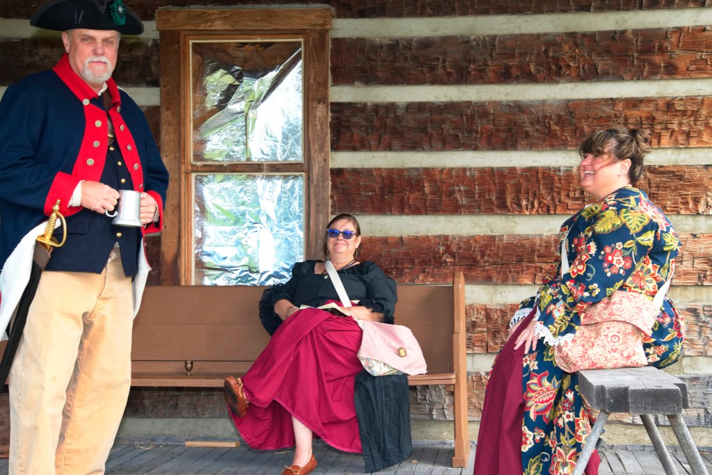 Three Colonial re-enactors gather on a long cabin's porch.