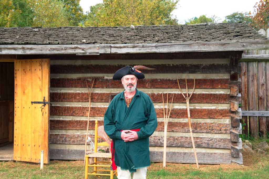 A costumed re-enactor in front of a log cabin.