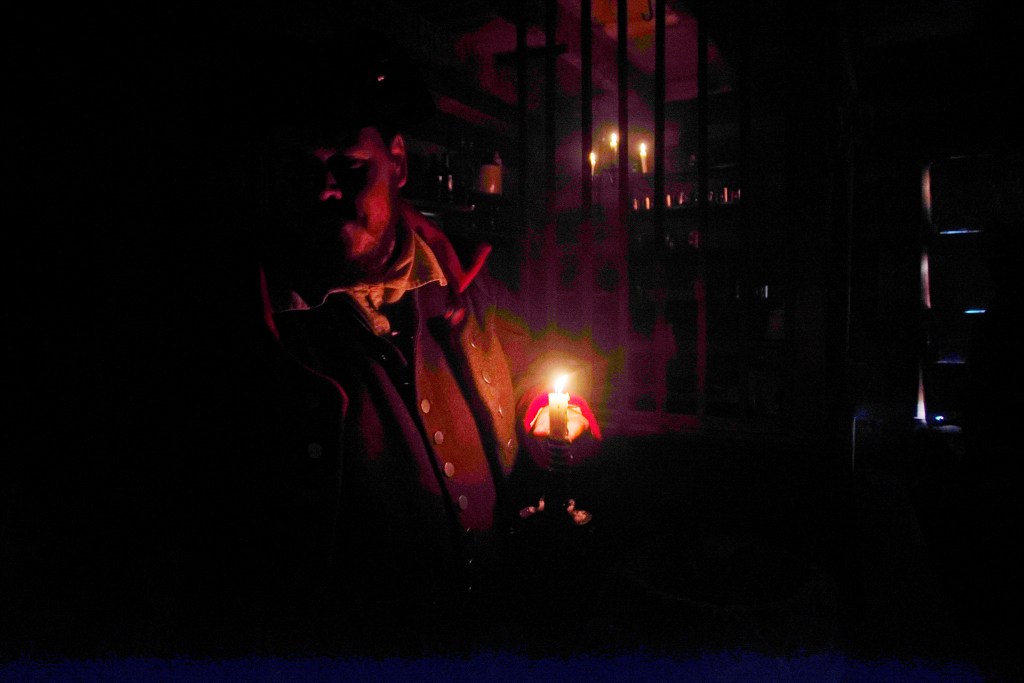 A man stands in a dark cabin with only one candle burning. 