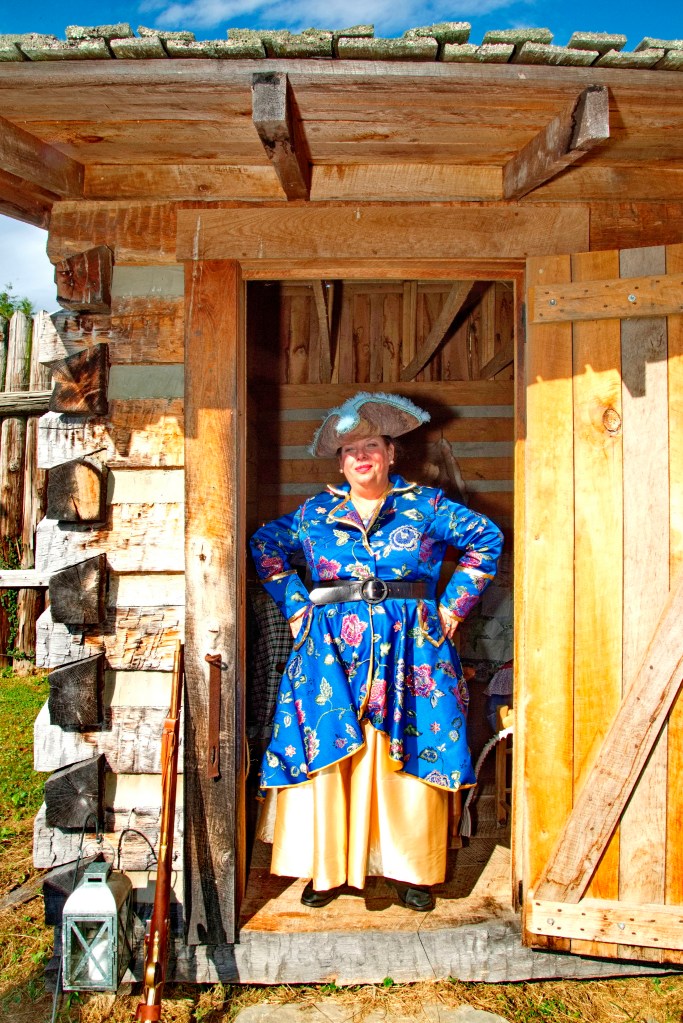 A costumed storyteller stands in the doorway of a log cabin. 