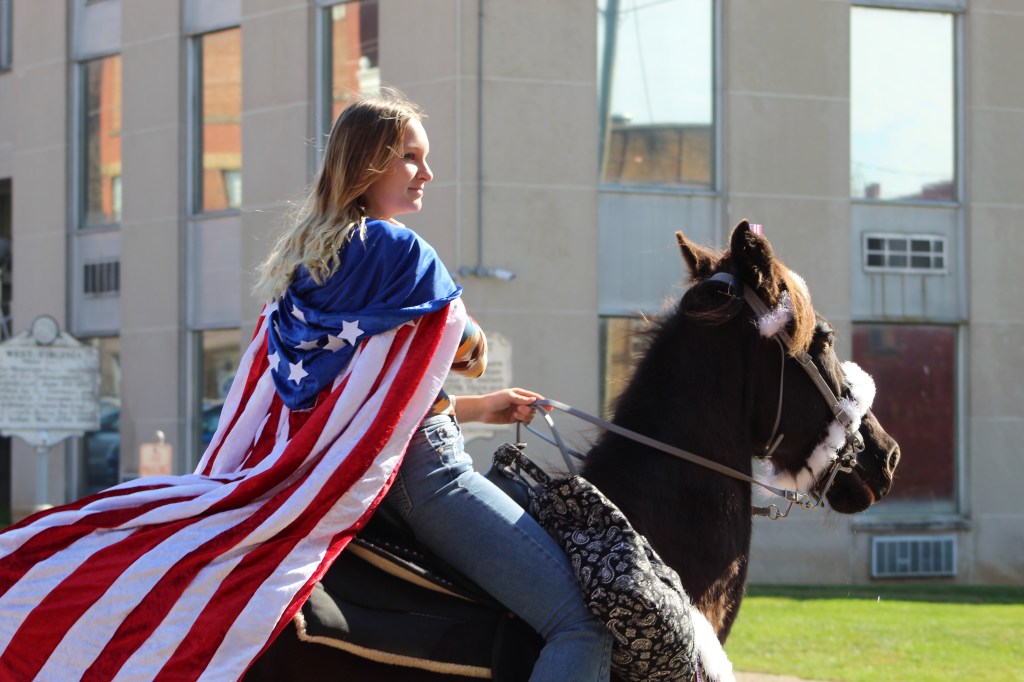 A young woman rides a horse with a patriotic flag on her back.