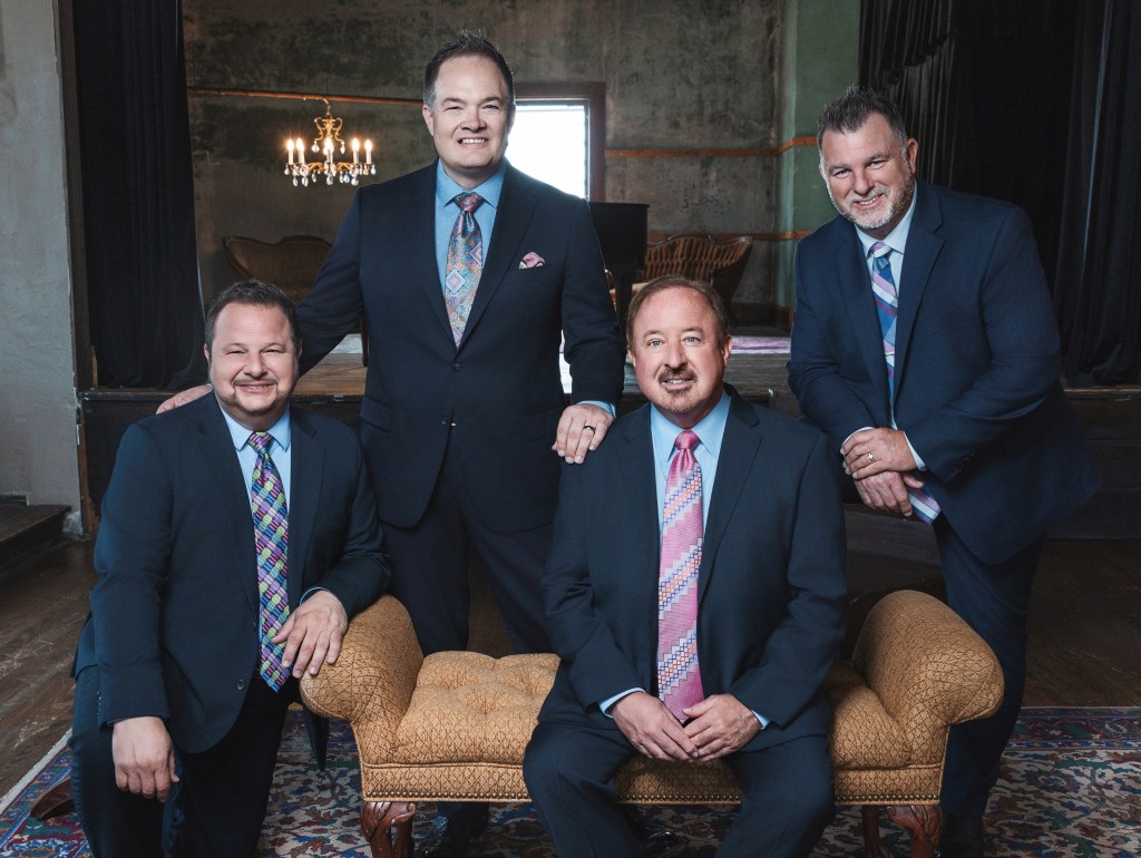 Pictured are four men who form the Southern Gospel quartet, The Guardians. The men pose inside a furnished lobby, wearing suits and ties, smiling directly at the camera. 